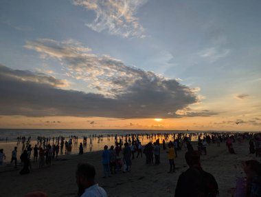 Gün batımında bir plajda toplanan kalabalık renkli gökyüzü ve yansıyan su akşam rahatlaması için sakin bir atmosfer yaratıyor.