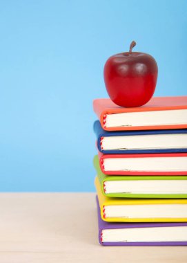 Parlak renkli ciltli kitaplar mavi arka planlı bir açık tahta masaya istiflenmiş. Yığılmış kitapların üstünde kırmızı elma. 