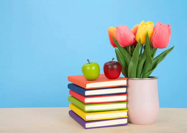 Bright colorful bound books stacked on a light wood table with blue background. Red apple on top of stacked books. Pink vase with tulip flowers sitting next to books.