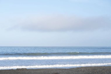 Kuzey Kaliforniya 'da kumlu sahil kenarında dalgalar kopuyor, açık mavi gökyüzünde tek uzun bulut..