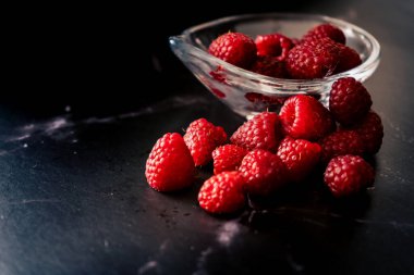High view of a Serving of raspberries in a small clear bowl on a black granite surface. Dark Food, horizontal orientation, space for text.
