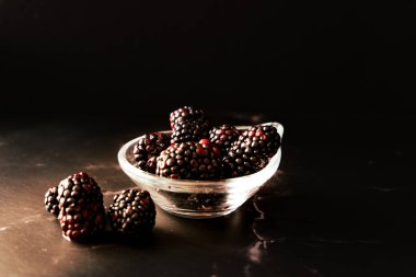 High view of a Serving of real Blackberries in a small clear bowl on a black granite surface. Horizontal orientation, space for text. Dark food.