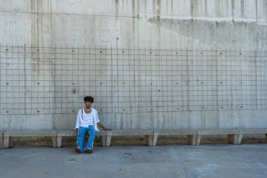 Kişisel stili olan yakışıklı genç bir Latin adam büyük bir beton duvarın önünde bankta oturuyor..