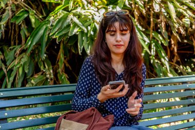 Arka planda yemyeşil yemyeşil bir bankta otururken akıllı telefon kullanan odaklanmış genç bir kadın.