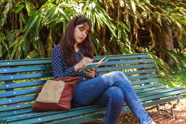Üniversite öğrencisi güzel güneşli bir günde bir bankta otururken notlar alıyor.