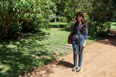 Güneşli bir parkta cep telefonu kullanan şık bir kadın iletişim ve boş vakitlerin tadını çıkarıyor.