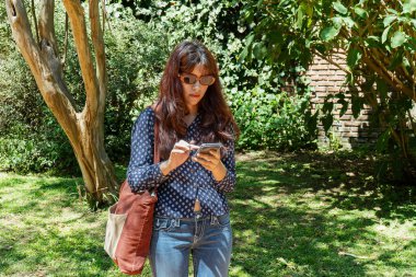 Cep telefonu uygulaması kullanan bir turist şehir parkında bankta oturuyor ve yaz gününün tadını çıkarıyor.