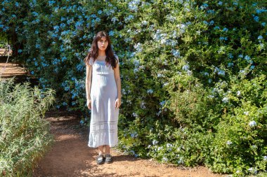 Hamile kadın mavi çiçeklerle dolu bir bahçede güneşli bir günün tadını çıkarıyor.