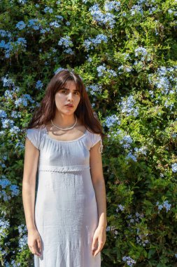 Çiçekli bir çitin önünde beyaz bir elbise ve inci kolye giyerek poz veren manken.