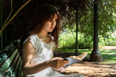 Güneşli bir parkın gölgeli bir alanında edebiyat ve doğaya dalmış bir kitaptan zevk alan bir kız.