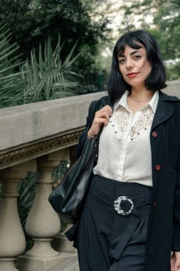 Kendine güveni tam bir iş kadını klasik bir parkta yürüyor, bir çanta taşıyor ve şık kıyafetler giyiyor.