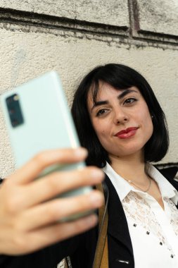 Zeki telefonuyla beton duvarın önünde selfie çekerken gülümseyen zarif İspanyol iş kadını.