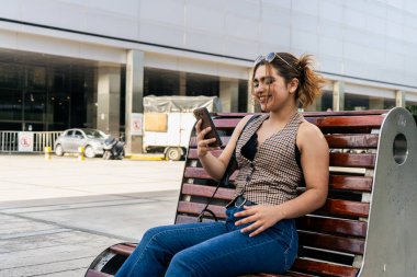 Genç iş kadını akıllı telefon kullanarak gülümsüyor ve ofis binasının dışındaki bankta dinleniyor.