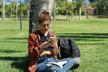 Üniversite öğrencisi parktaki ağacın altında sırt çantası ve kitapla akıllı telefon kullanıyor.