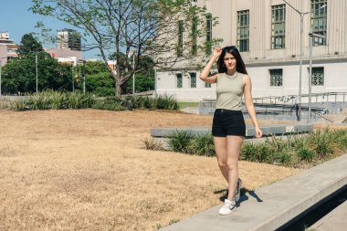 Şık bir genç kadın şehir parkında geziyor, güneşli havanın tadını çıkarıyor.