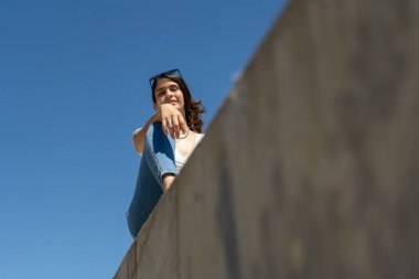 Şık, genç İspanyol bir kadın güneşli bir günün tadını çıkarıyor, beton bir duvarda canlı mavi bir gökyüzüne karşı oturuyor.