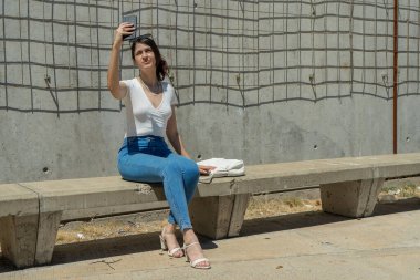 Genç İspanyol kadın beton bir bankta otururken akıllı telefonuyla selfie çekiyor.