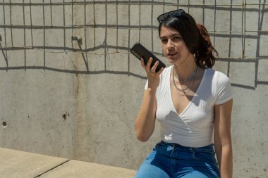 Genç Latin bir kadın şehir ortamında otururken akıllı telefonunu kullanarak sesli mesaj gönderiyor.