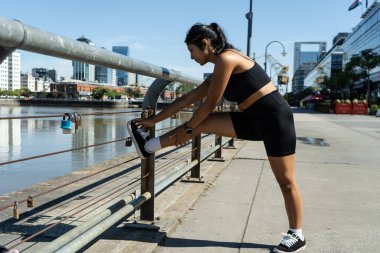 Genç Latin sporcu kadın Puerto madero nehrinin kıyısında bacaklarını esnetiyor.