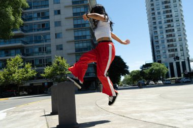 Kırmızı ve beyaz çizgili pantolon ve beyaz tişört giyen genç Latin bir kadın şehir ortamında beton bir bloğun üzerinden atlıyor.