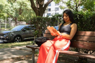 Genç Latin sporcu kadın, spor salonu ve edebiyatı birleştirerek parkta otururken kitap okumaktan zevk alıyor.