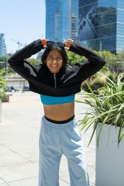 Gülümseyen İspanyol sporcu kadın modern şehir ortamında kafasına güneş gözlüğü takıyor, antrenmanına hazır.