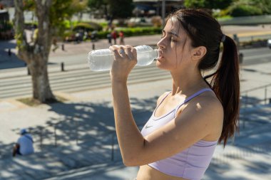 Genç sporcu kadın dışarıda çalıştıktan sonra plastik şişeden su içiyor, fiziksel aktiviteden sonra ferahlatıyor ve susuzluğunu gideriyor.