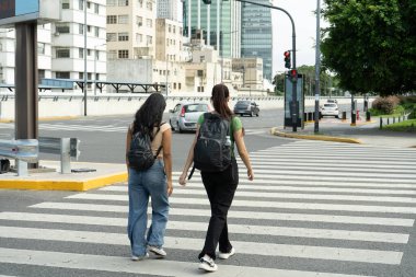 İki kız öğrenci kalabalık bir şehir ortamında yaya geçidinde yürüyorlar.