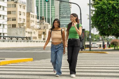 İki kız öğrenci büyük bir şehirde yaya geçidinde yürüyor ve konuşuyor.