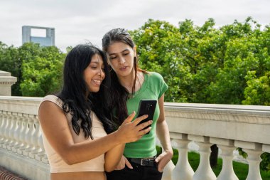 İki genç kadın gülümsüyor ve şehir parkında akıllı bir telefona bakıyorlar.