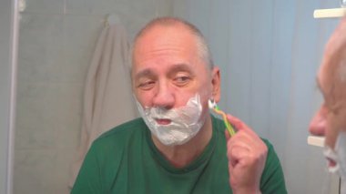 Yetişkin bir adam sakalını kesmek için tıraş köpüğüyle yüzünü jiletle kapatmış..