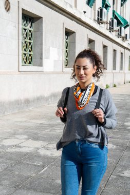 Şık bir genç kadın, elinde bir sırt çantası ve şık bir eşarp ile bir binanın yanında yürüyor.