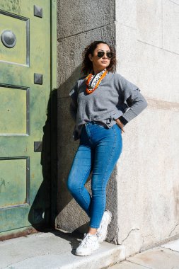 Güneş gözlüğü takan şık bir kadın ve şehir sokaklarında duvara yaslanmış bir eşarp.