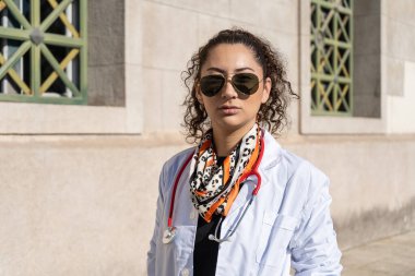 Hastanenin önünde duran güneş gözlüğü ve steteskop takan ciddi bir kadın doktorun portresi.