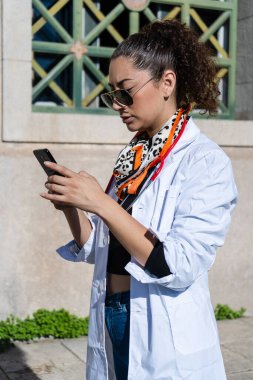 Beyaz önlük ve güneş gözlüğü takan genç bayan doktor dışarıda dururken cep telefonu kullanıyor.