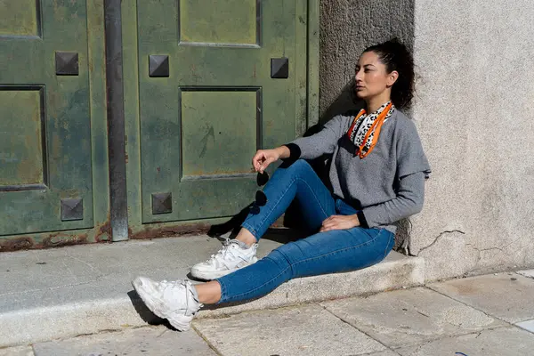 Eski bir binanın önünde oturan, elinde güneş gözlüğü olan, güneşli bir günün tadını çıkaran modaya uygun genç bir kadın.