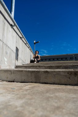 Sporcu kadın şehirde beton basamaklarda mola veriyor, serinletici bir içkinin tadını çıkarıyor.