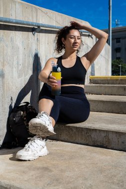 Genç bir kadın idmana ara veriyor, beton merdivenlerde oturuyor ve elinde bir su şişesi tutuyor.