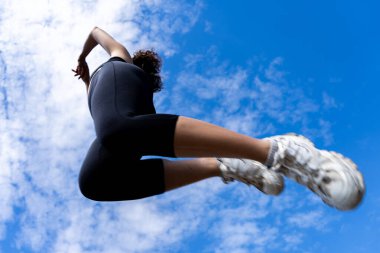 Mavi bulutlu gökyüzünün altında yürüyen siyah spor giysili genç bir kadının alçak açılı görüntüsü.