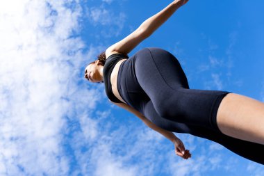 Mavi bulutlu gökyüzünün altında yürüyen siyah spor giysili genç bir kadının alçak açılı görüntüsü.