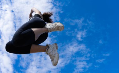 Sporcu kadın mavi gökyüzü ve bulutlardan oluşan bir zemine güçlü bir atlayış yapıyor, atletizm ve kararlılık sergiliyor.