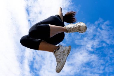 Sporcu kadın mavi gökyüzü ve bulutlardan oluşan bir zemine güçlü bir atlayış yapıyor, atletizm ve kararlılık sergiliyor.