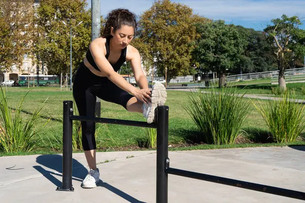 Parkta korkuluk kullanarak bacaklarını açan sporcu kadın güneşli bir günün tadını çıkarıyor.