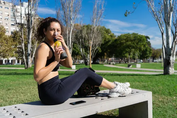 Parktaki antrenmandan sonra yedekte dinlenirken izotonik içki içen genç bir kadın.