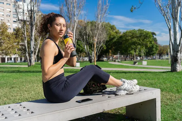 Parktaki antrenmandan sonra yedekte dinlenirken izotonik içki içen genç bir kadın.