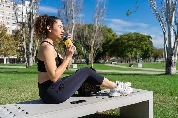 Parktaki antrenmandan sonra yedekte dinlenirken izotonik içki içen genç bir kadın.