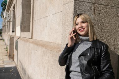 Güneşli bir günde duvara yaslanırken akıllı telefondan konuşan modaya uygun genç bir kadın.