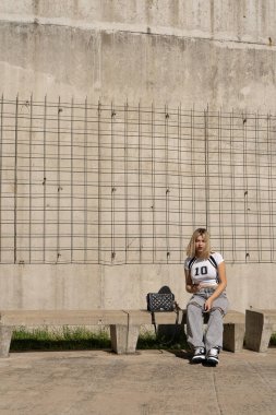 Şehir ortamında beton bir bankta otururken akıllı telefonunu kullanan zarif genç bir kadın. Arkaplan olarak tel örgüler ve beton duvarlar var.