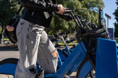 Üniversite öğrencisi yırtık kot pantolon giyiyor mavi bir bisikletin yanında duruyor bir üniversite kampüsünde bisikleti paylaşıyor.