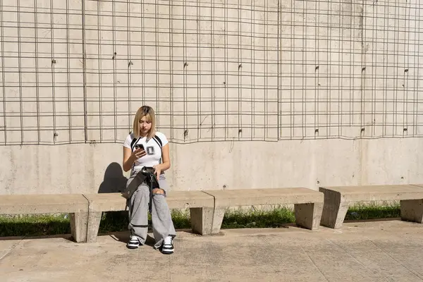 Genç bir öğrenci, beton bir duvarın önünde metal ızgaralı bir bankta otururken çantasını açıyor.